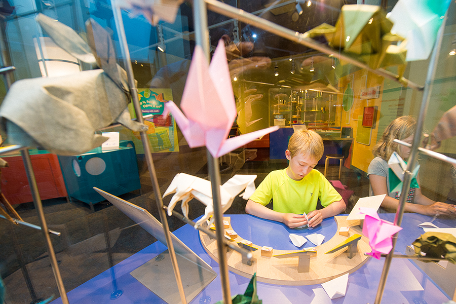 A boy folds an origami shape at a table surrounded by other origami creations.